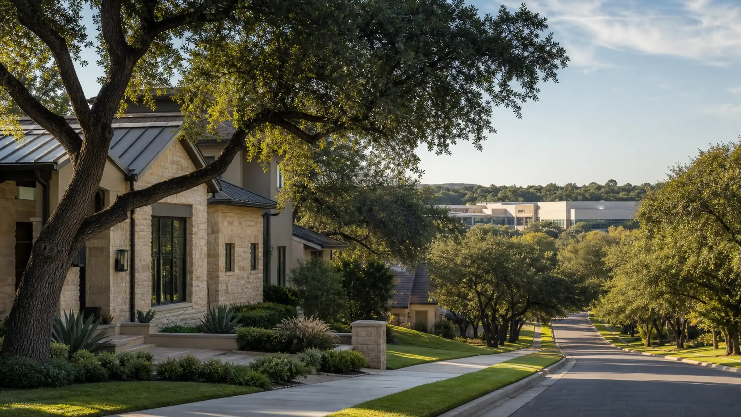 A stone-clad home with a standing-seam metal roof and mature live oak trees along a sidewalk-lined residential street in the San Antonio area